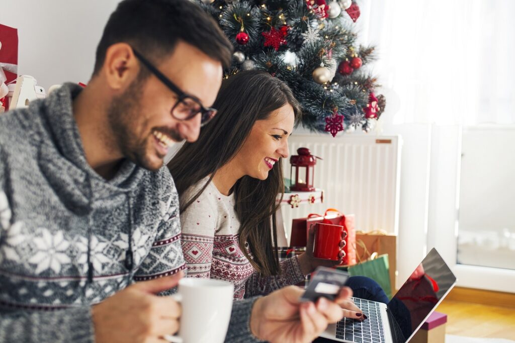 Moteris ir vyras sėdi prie Kalėdų eglutės ir žiūri į nešiojamąjį kompiuterį.