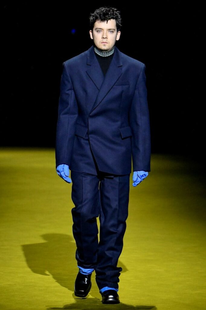 Modelis, dėvintis tamsiai mėlyną kostiumą ir juodus batus