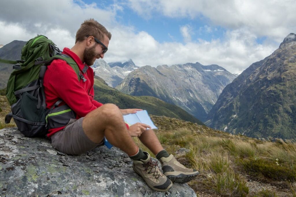 Kalnuose ant akmens sėdintis vyras, apsirengęs raudonu sportiniu džemperiu, rudais sportiniais šortais.