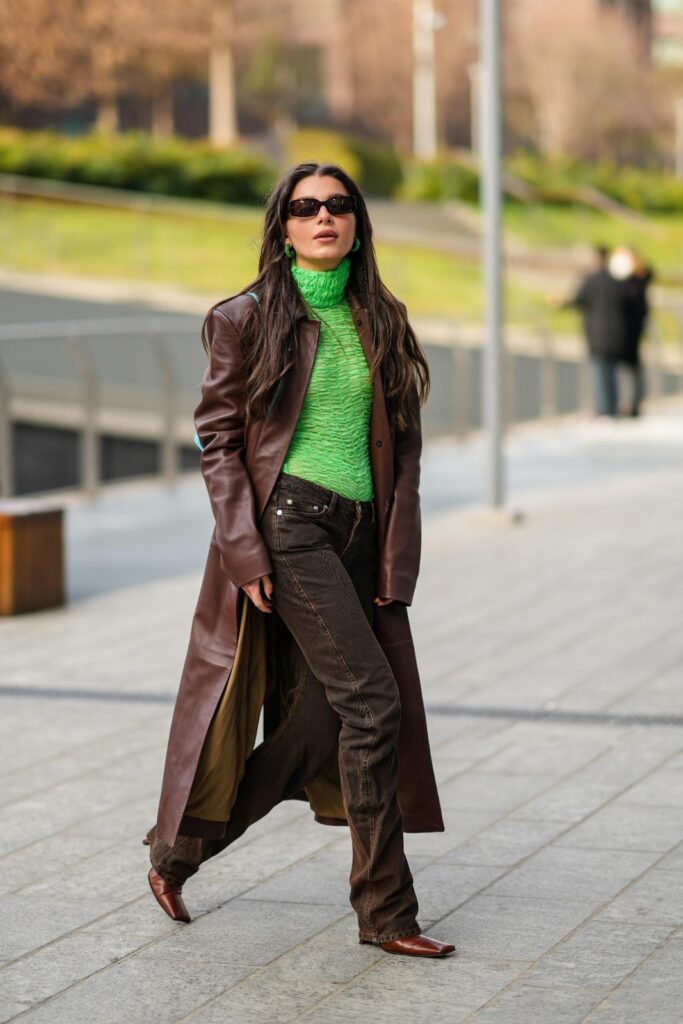 Moteris, dėvinti žalią golfą, rudą odinį paltą, rudus džinsus ir rudus aulinukus